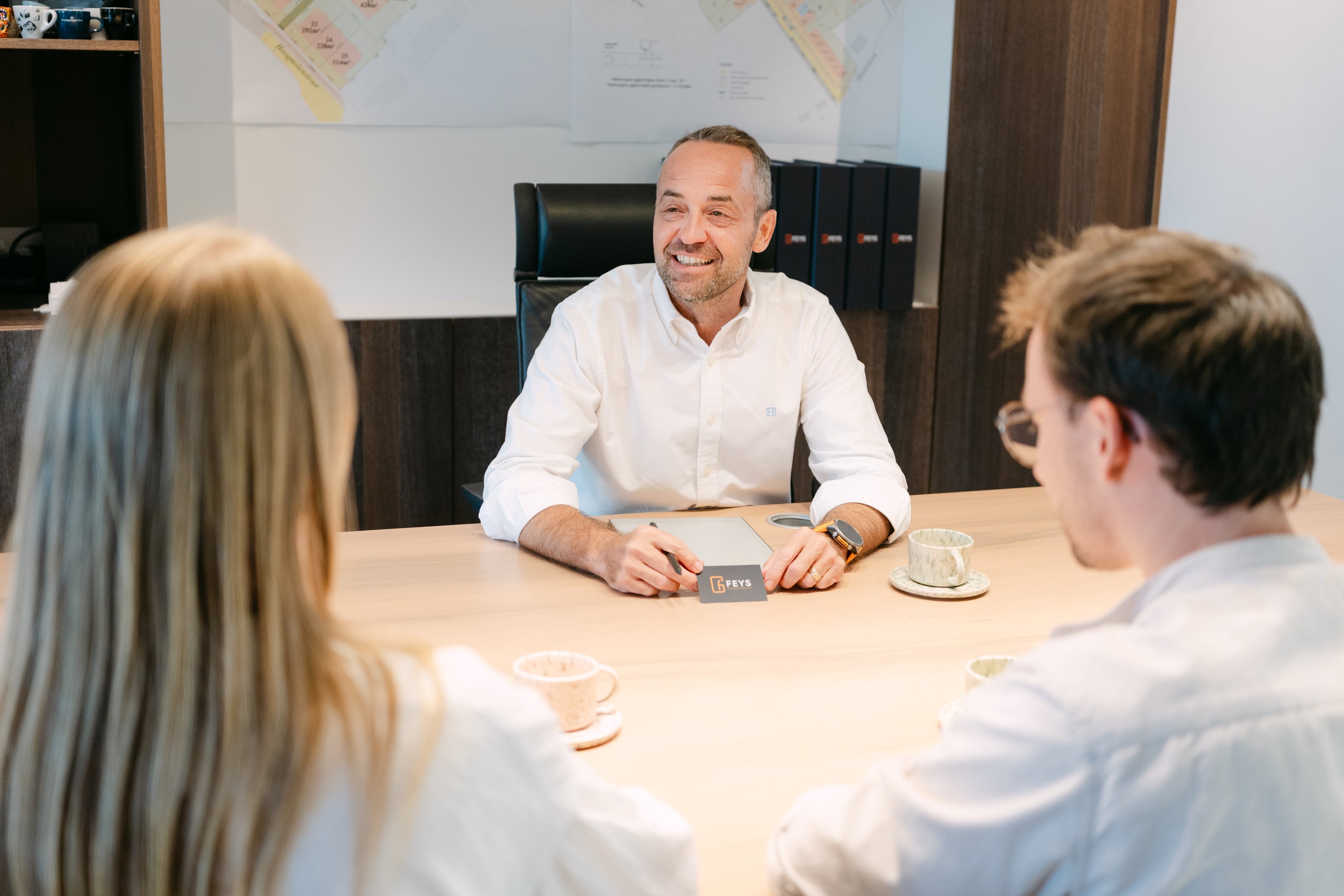 Man in wit overhemd zit aan houten tafel in gesprek met twee personen in kantooromgeving